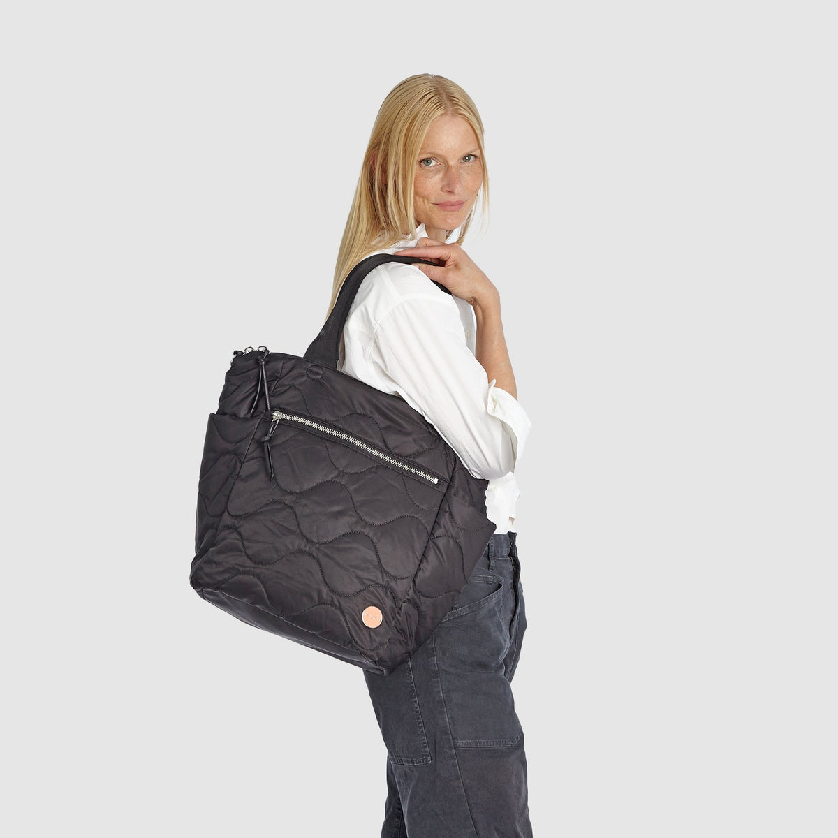 Woman standing next to a black ranger quilted tote bag and silver suitcase on a white background