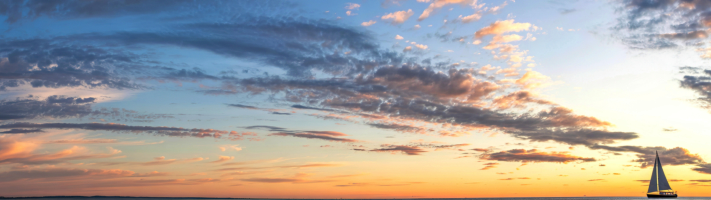 sailboat appears small against blue, pink and gold clouds at sunset