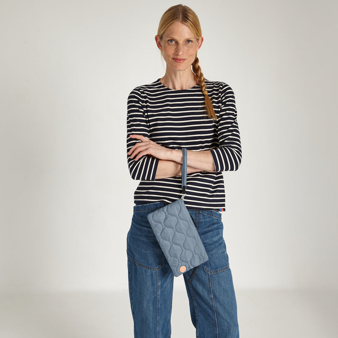 shortyLOVE cleo wristlet clutch in steel blue; on model's wrist in striped shirt and jeans against white background.