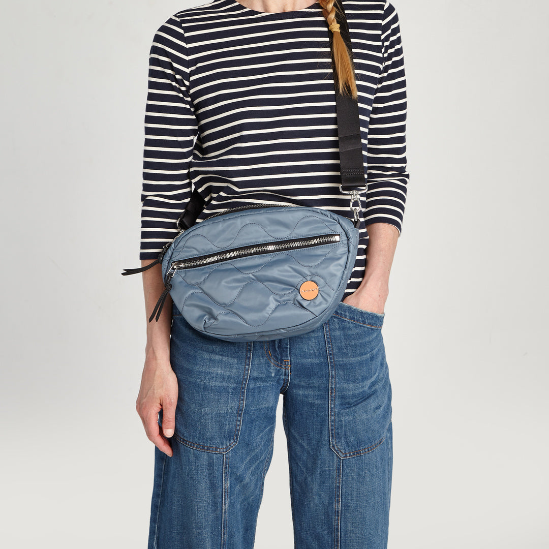 shortyLOVE cruiser medium crossbody bag in steel blue; on model in striped shirt and jeans against white background.