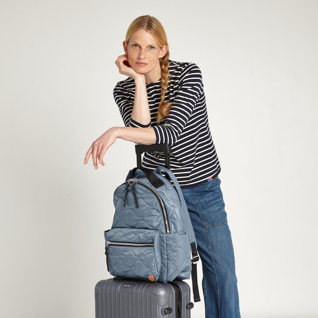 shortyLOVE tate backpack in steel blue; on luggage with model in striped shirt and jeans against white background.