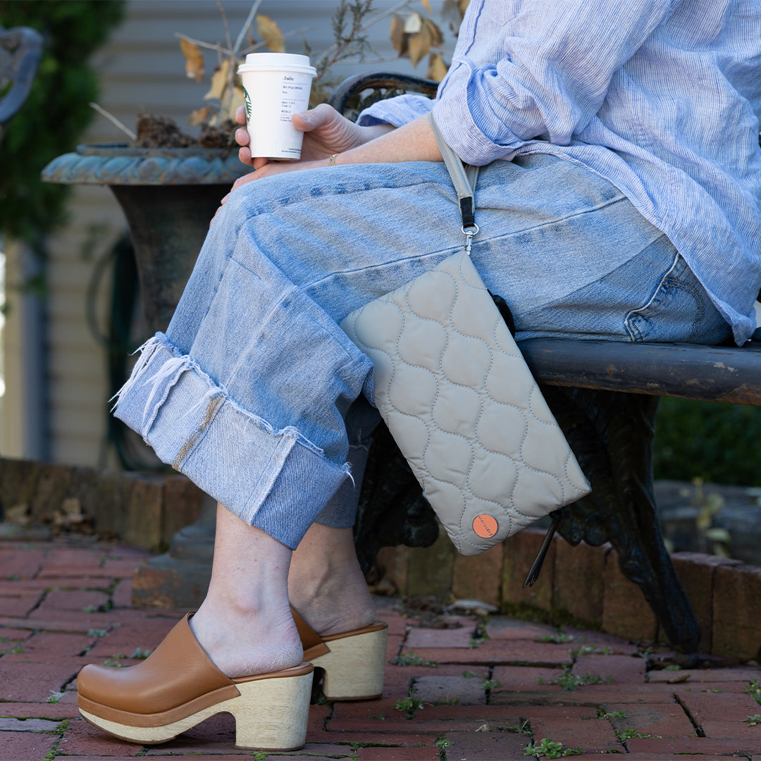 Person sitting on a bench wearing denim pants and brown clogs, holding a coffee cup and a grey cleo quilted clutch.