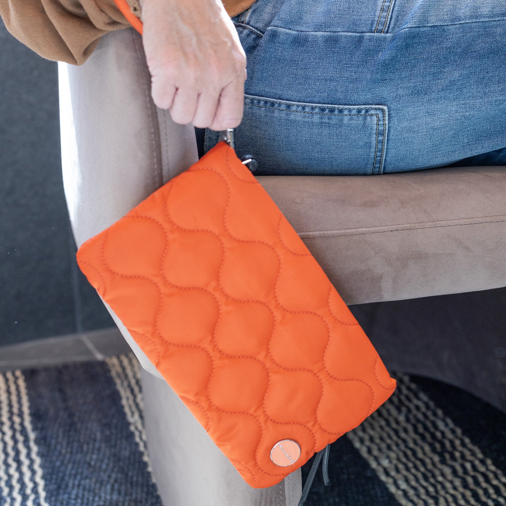 Person holding an orange quilted cleo clutch bag on a carpeted floor.