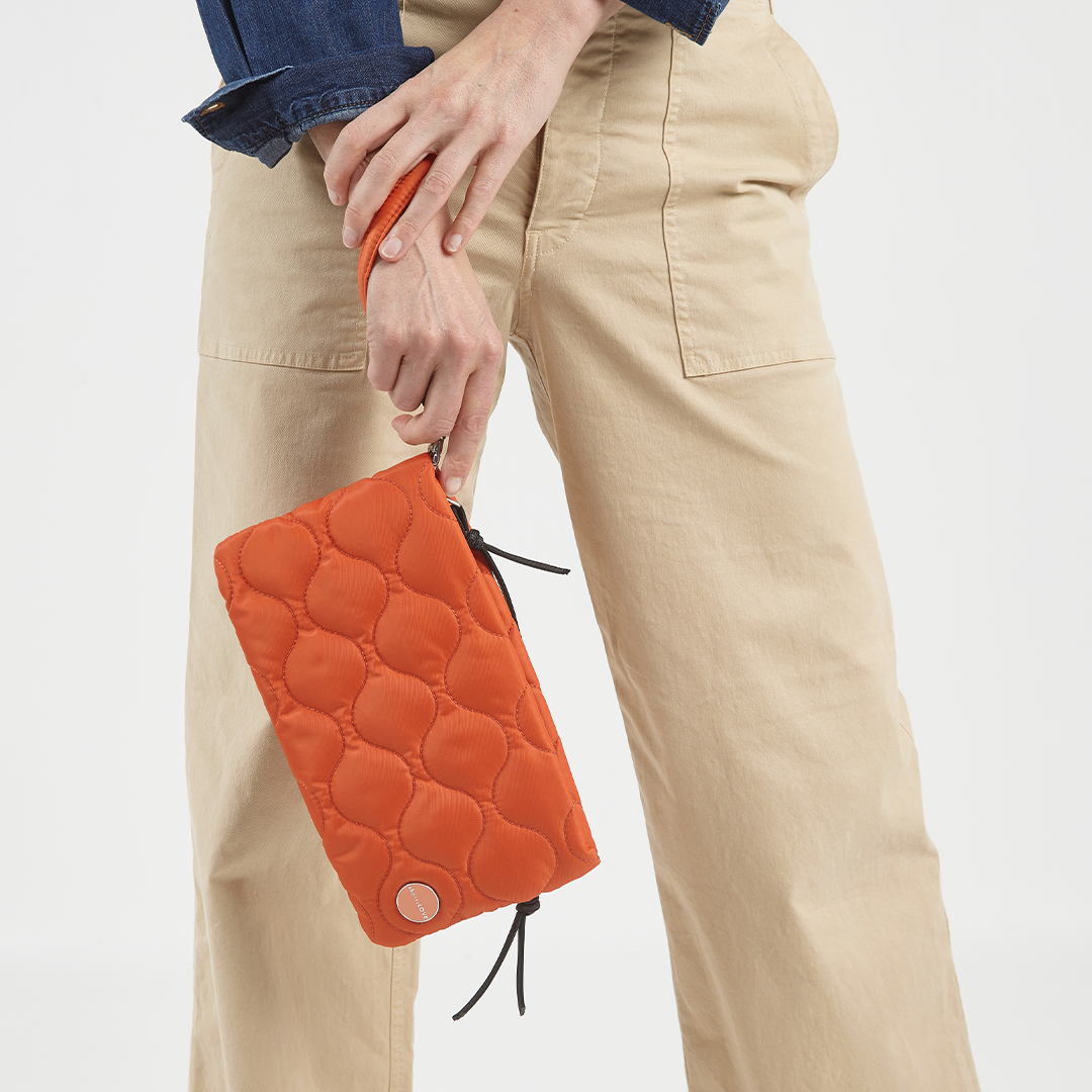 Person holding an orange quilted cleo clutch bag with beige pants and a blue jacket.