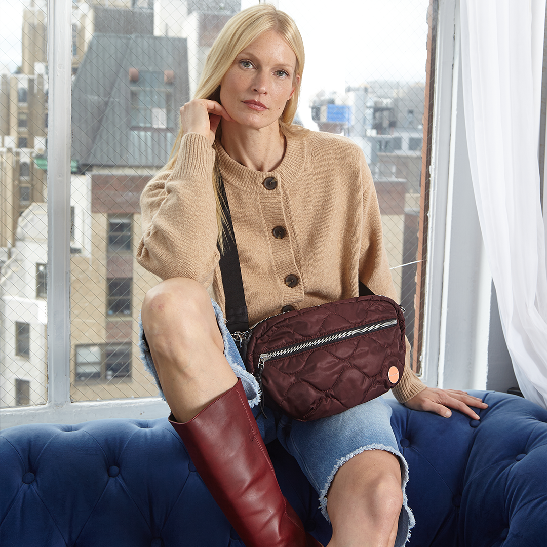 Woman sitting on a blue couch holding a bordeaux quilted cruiser handbag with a cityscape in the background.