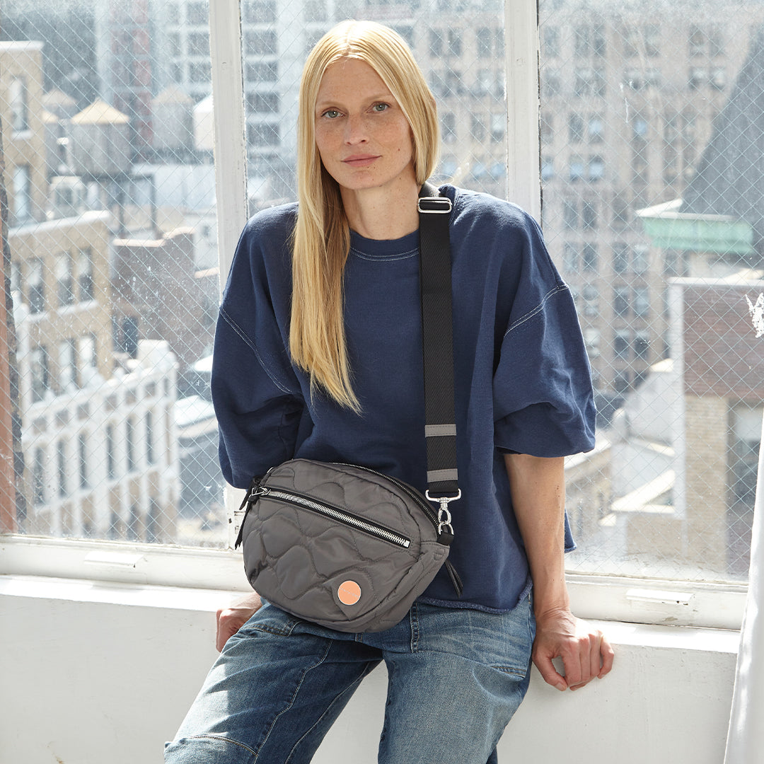 Person wearing a blue oversized shirt and jeans with a pewter quilted cruiser crossbody bag, standing in front of a window with cityscape view.