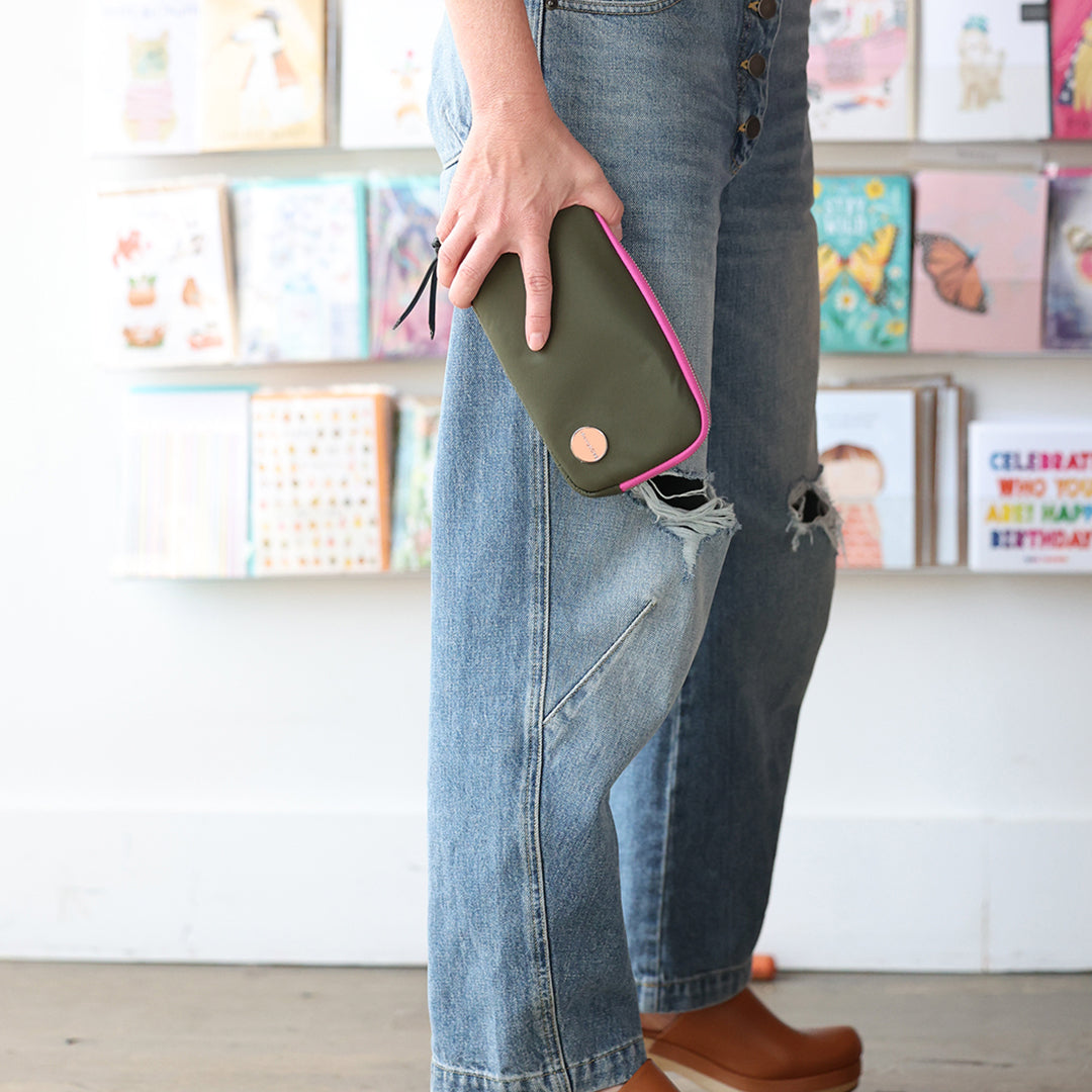 Person holding an army green and pink jetty wallet/clutch with a blurred background of books and decorations