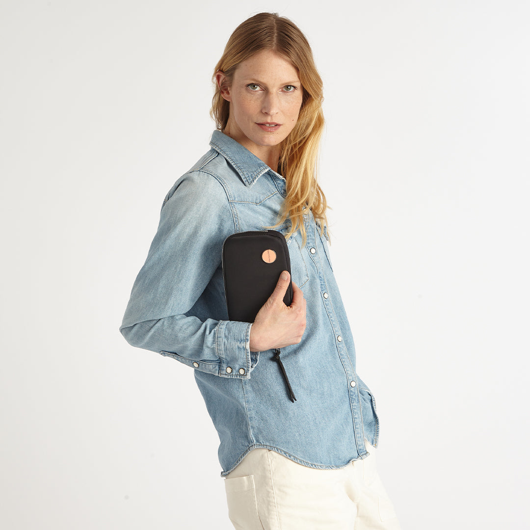 Woman wearing a light blue denim shirt holding a black jetty wallet/clutch with a brand logo.