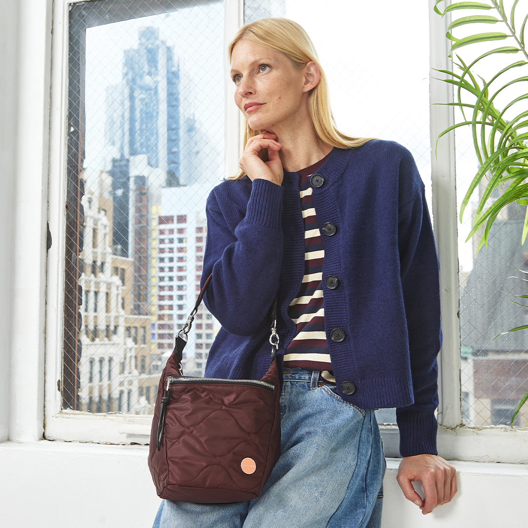 Woman in a blue cardigan with a striped shirt and jeans, holding a bordeaux marcel quilted crossbody bag, in front of a window with cityscape view.