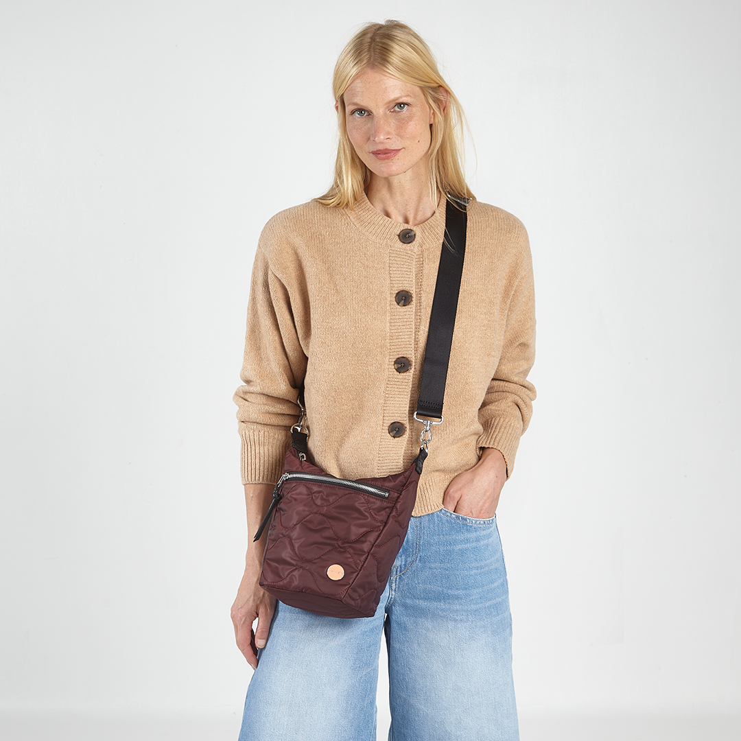 Woman wearing a beige cardigan and blue jeans, holding a bordeaux marcel quilted crossbody bag against a white background