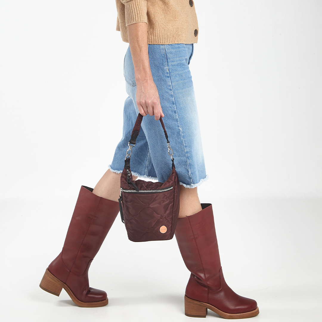 Person wearing maroon knee-high boots and holding a matching marcel quilted crossbody handbag on a white background