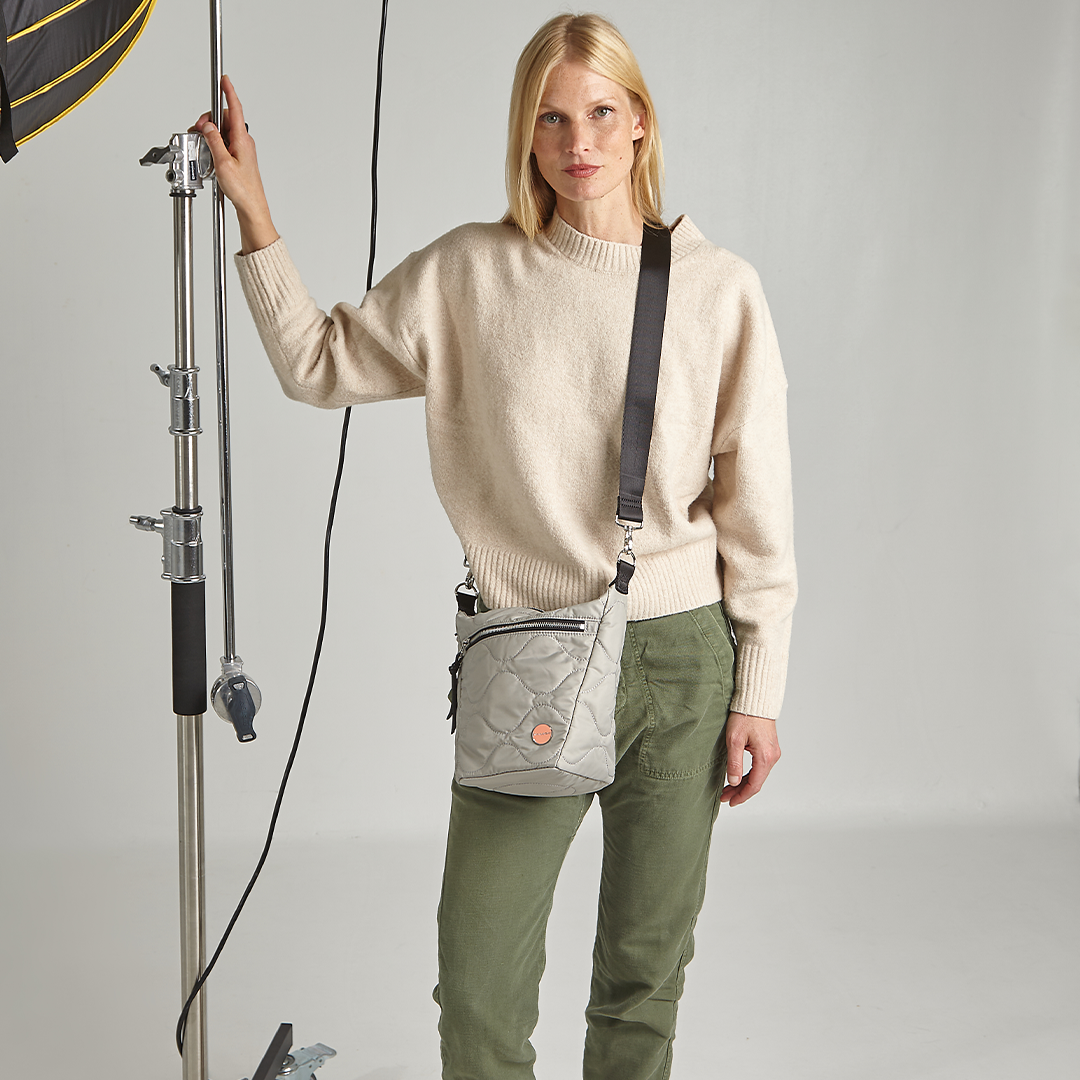 Woman wearing a beige sweater and green pants with a gray marcel quilted crossbody bag, standing in a studio setting.