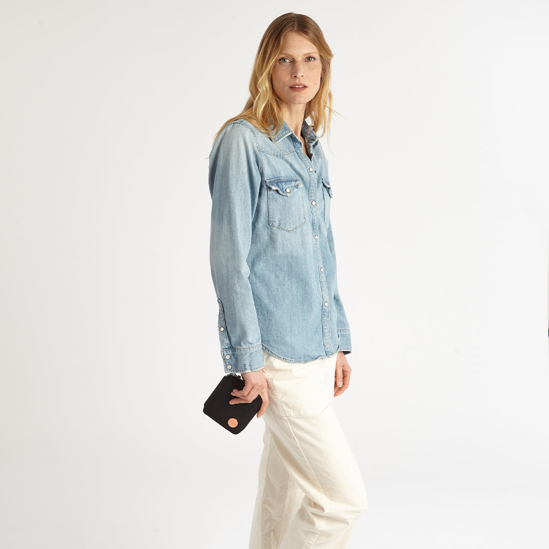 Woman wearing a light blue denim shirt and white pants holding a black merchant wallet on a plain background