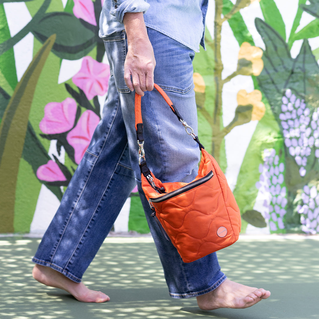 Person holding an orange marcel quilted crossbody bag with a colorful floral mural in the background