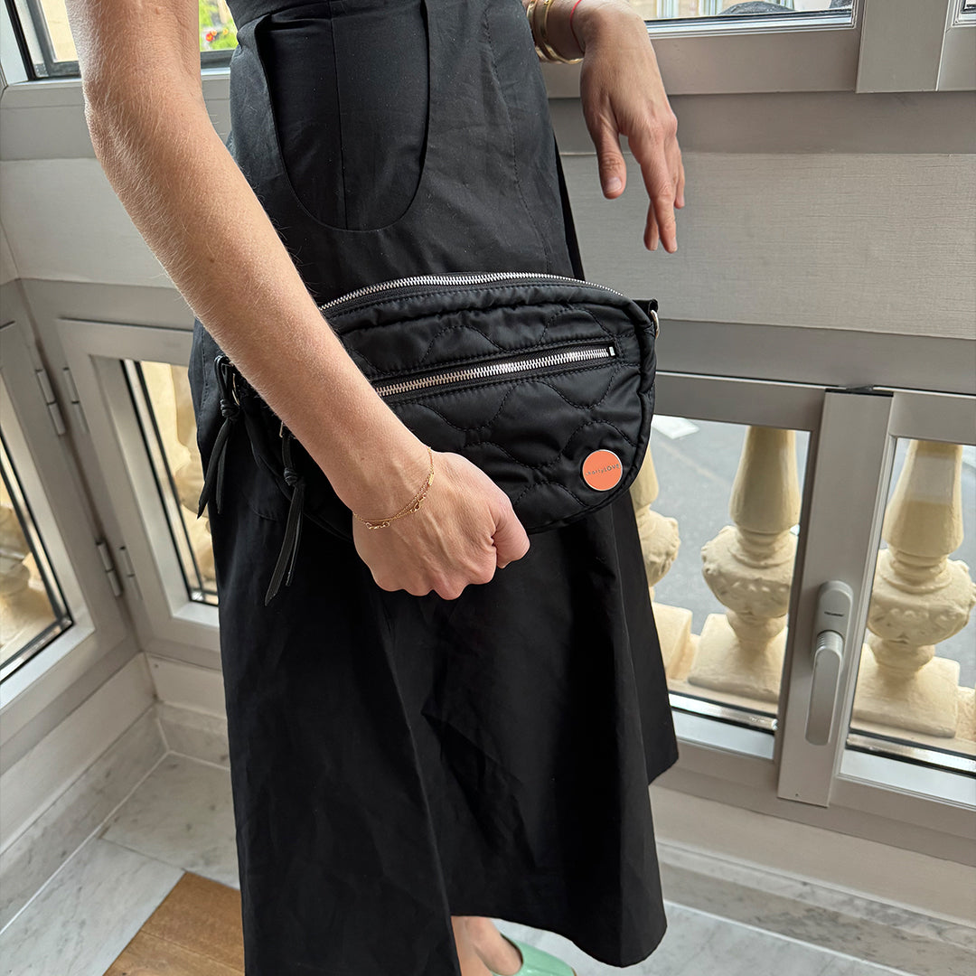 Person wearing a black dress with a black small cruiser quilted crossbody bag with a window background.