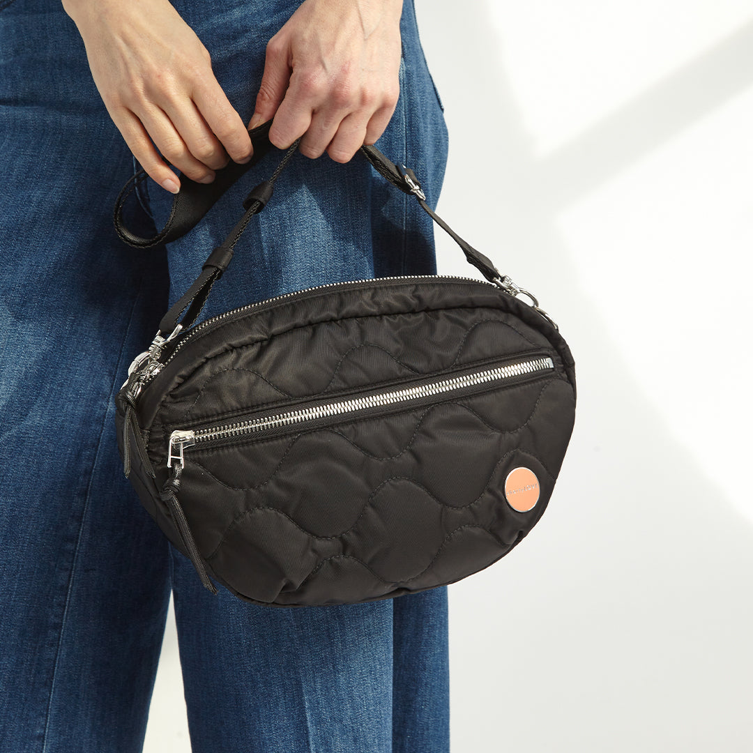 Black quilted small cruiser crossbody handbag held by a person wearing blue jeans on a white background