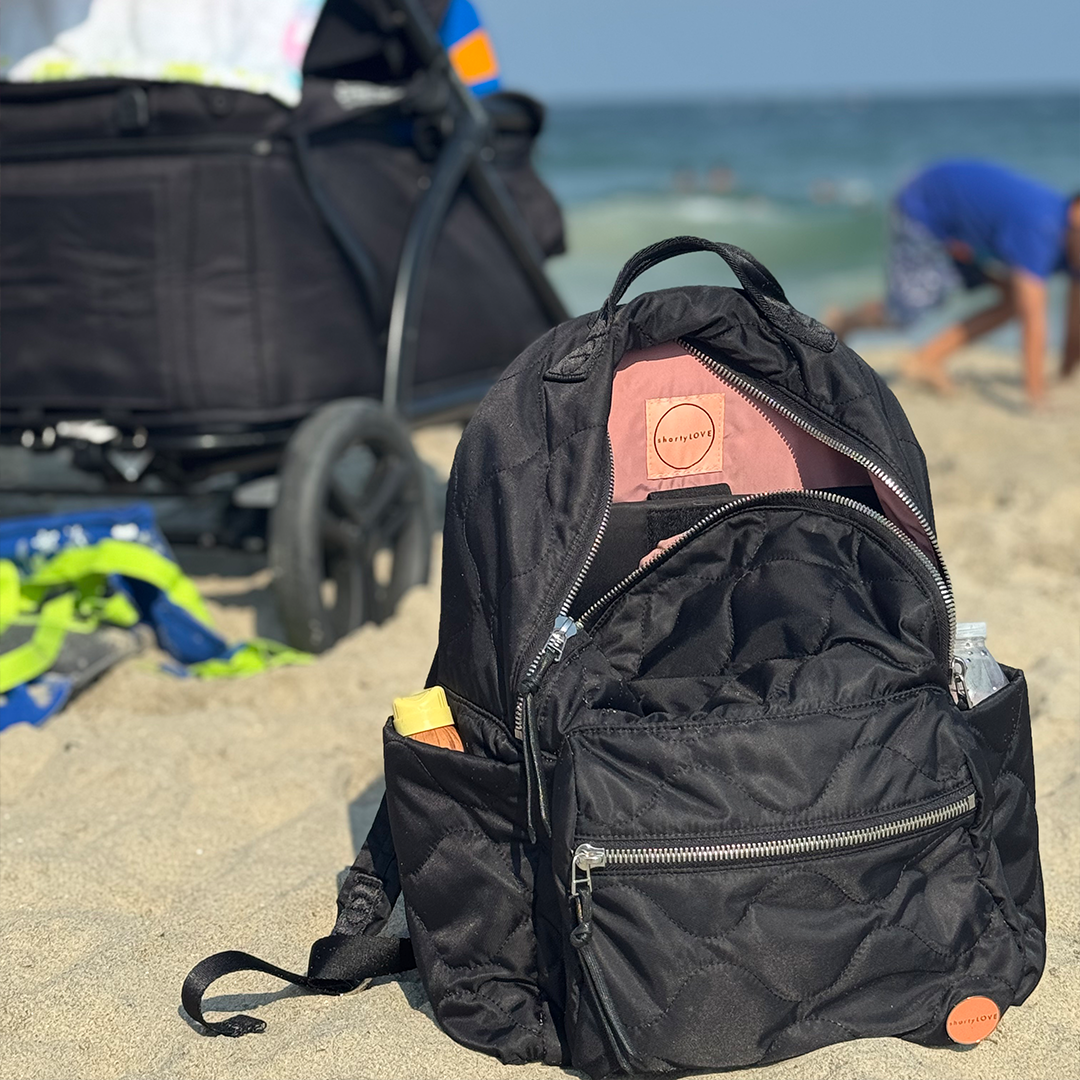 Black tate quilted backpack on a sandy beach with people and a stroller in the background.