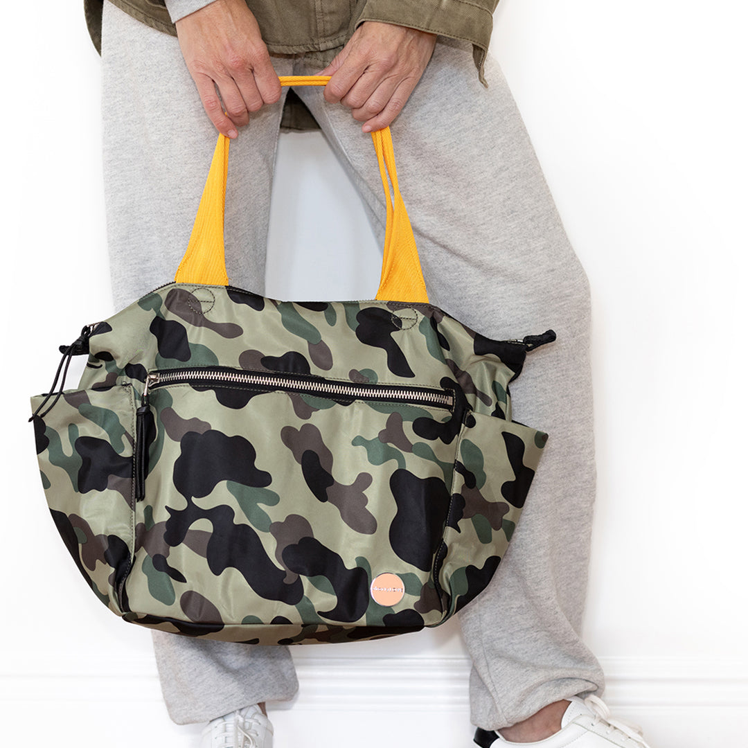 Camouflage-patterned tillie tote bag with yellow handles held by a person wearing gray pants.