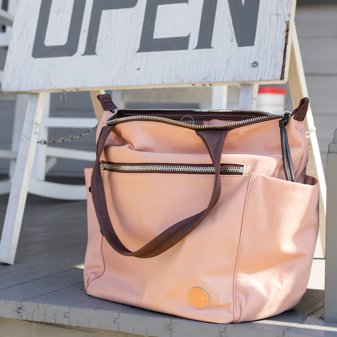 Pink/peach tillie tote bag with brown straps on a wooden surface, with 'OPEN' sign in the background.