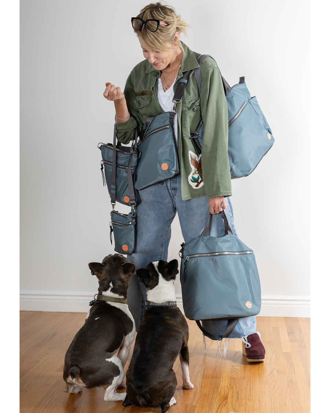 sloane in green jacket & jeans holding 5 styles of steel blue/charcoal bags looking down at Fred & Otis, her two dogs.