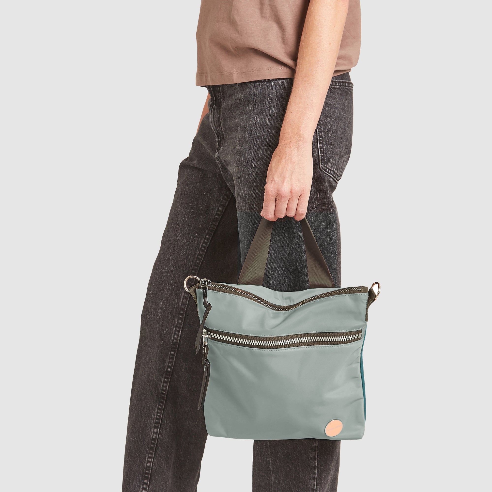 shortyLOVE amuse medium sized crossbody bag in slate/teal; in hand of model in blush rose top and black jeans against white background.