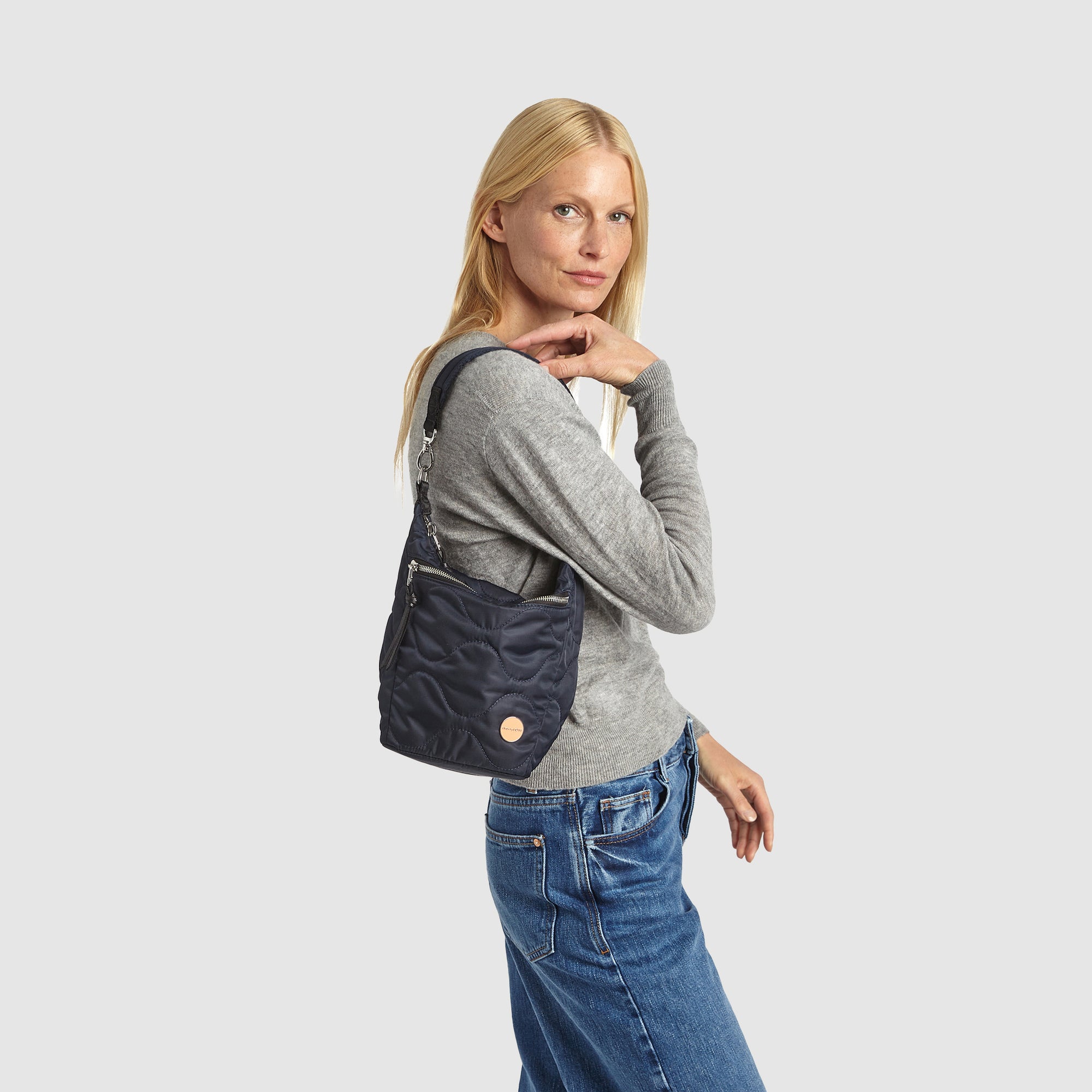 Woman wearing a gray sweater and blue jeans with a midnight blue marcel quilted crossbody bag on a white background