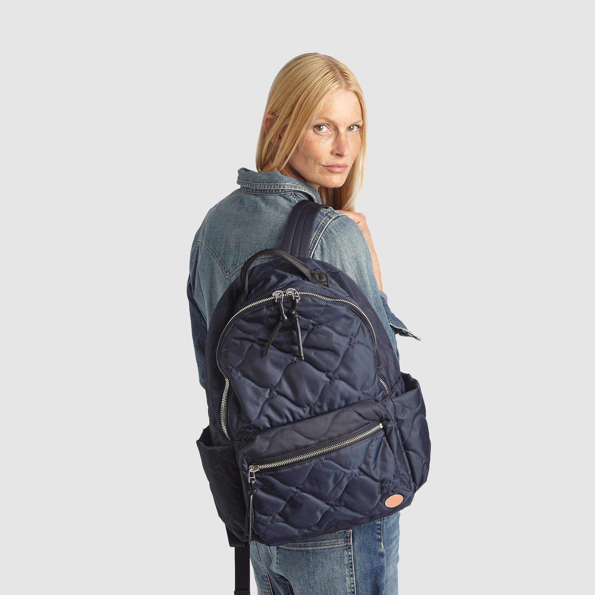 Woman in jean outfit holding a midnight blue tate quilted backpack on a white background