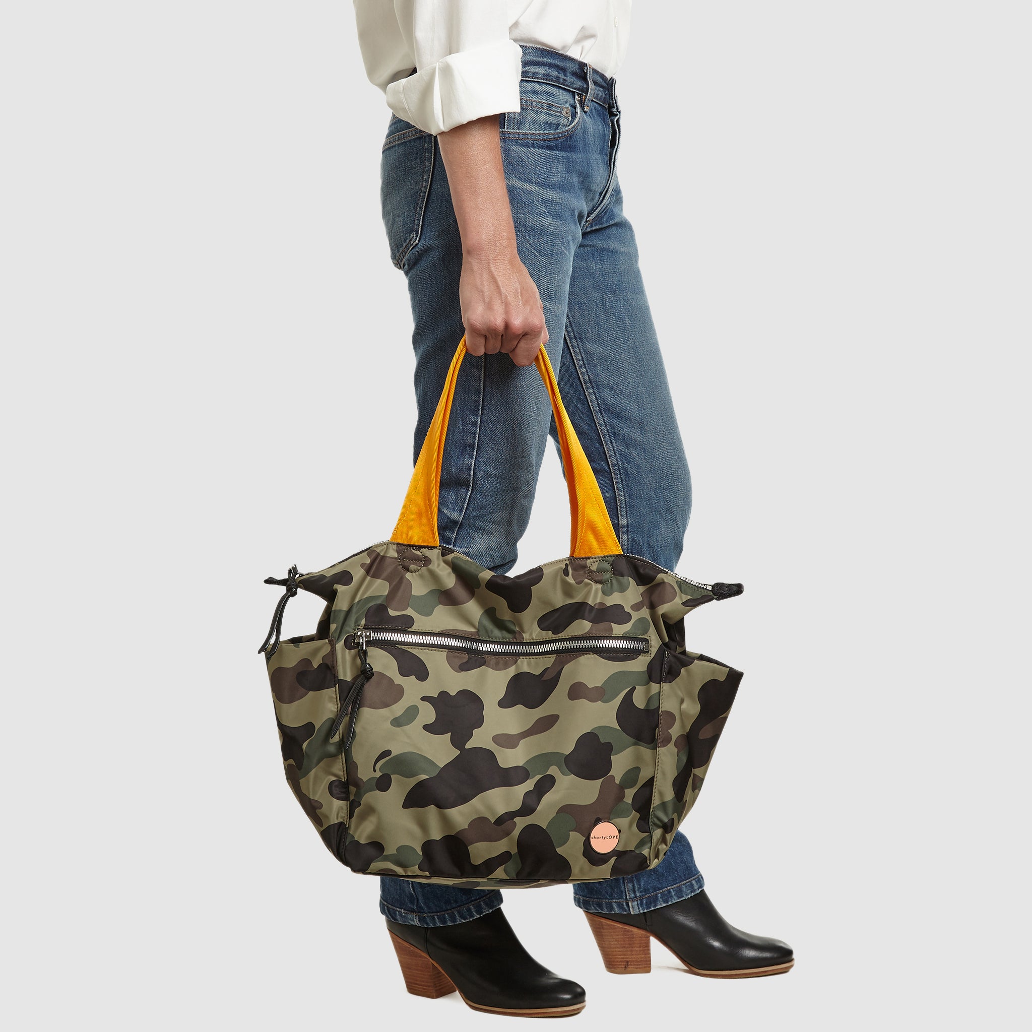 shortyLOVE tillie tote bag in green camo; hand held by model in white button down shirt and jeans against white background.