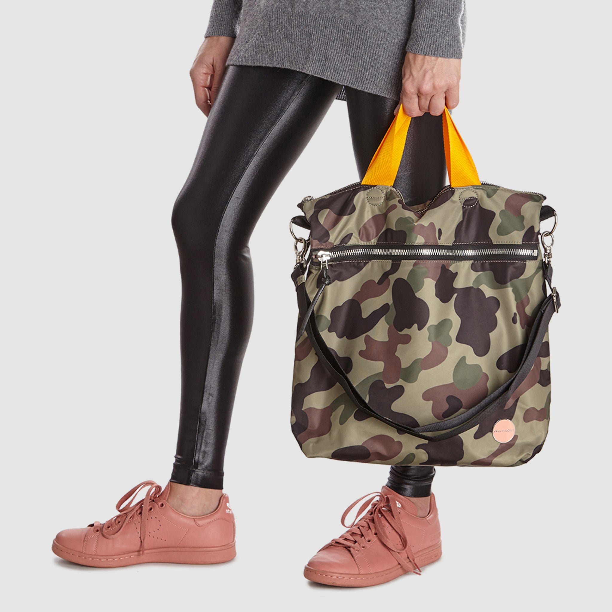 shortyLOVE wonder large crossbody/messenger bag in green camo; hand held by model in grey long sleeve sweater and black leggings against white background.