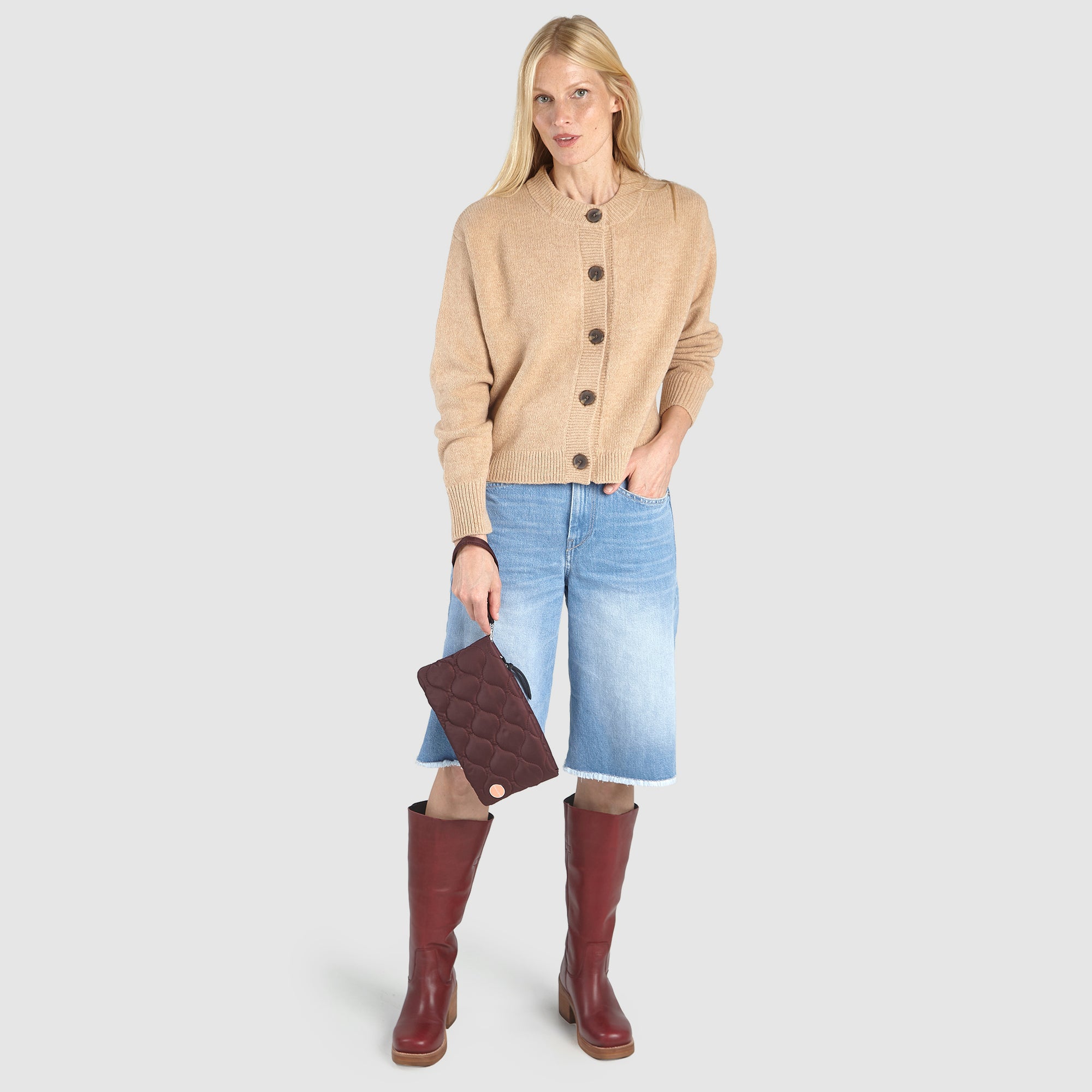 Person holding a bordeaux cleo quilted clutch with a beige sweater, blue denim skirt, and red boots on a white background