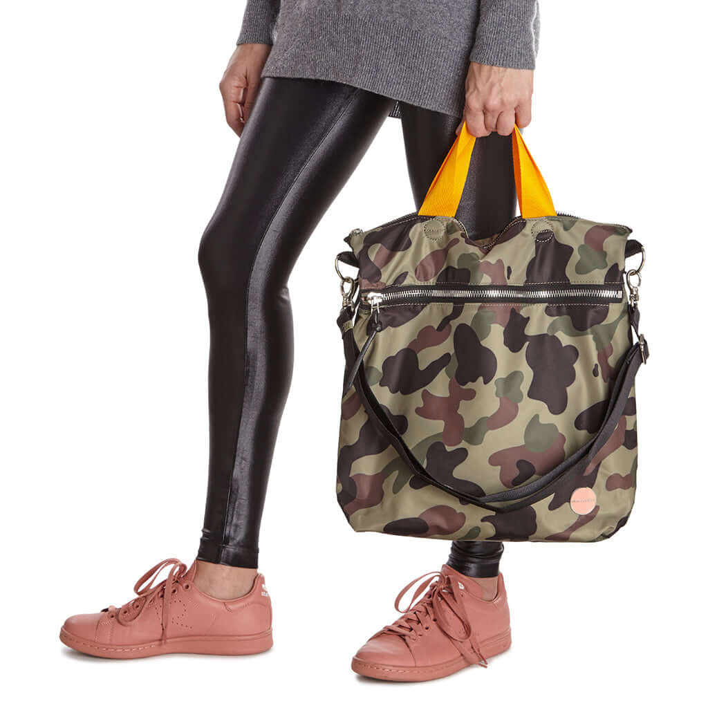 shortyLOVE wonder large crossbody/messenger bag in green camo; hand held by model in grey long sleeve sweater and black leggings against white background.