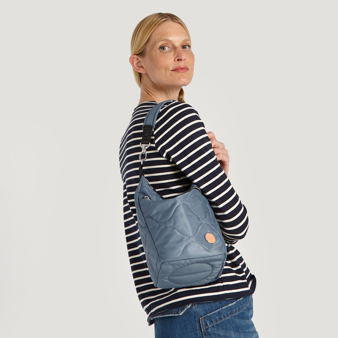 shortyLOVE marcel crossbody bucket bag in steel blue; on model in striped shirt and jeans against white background.