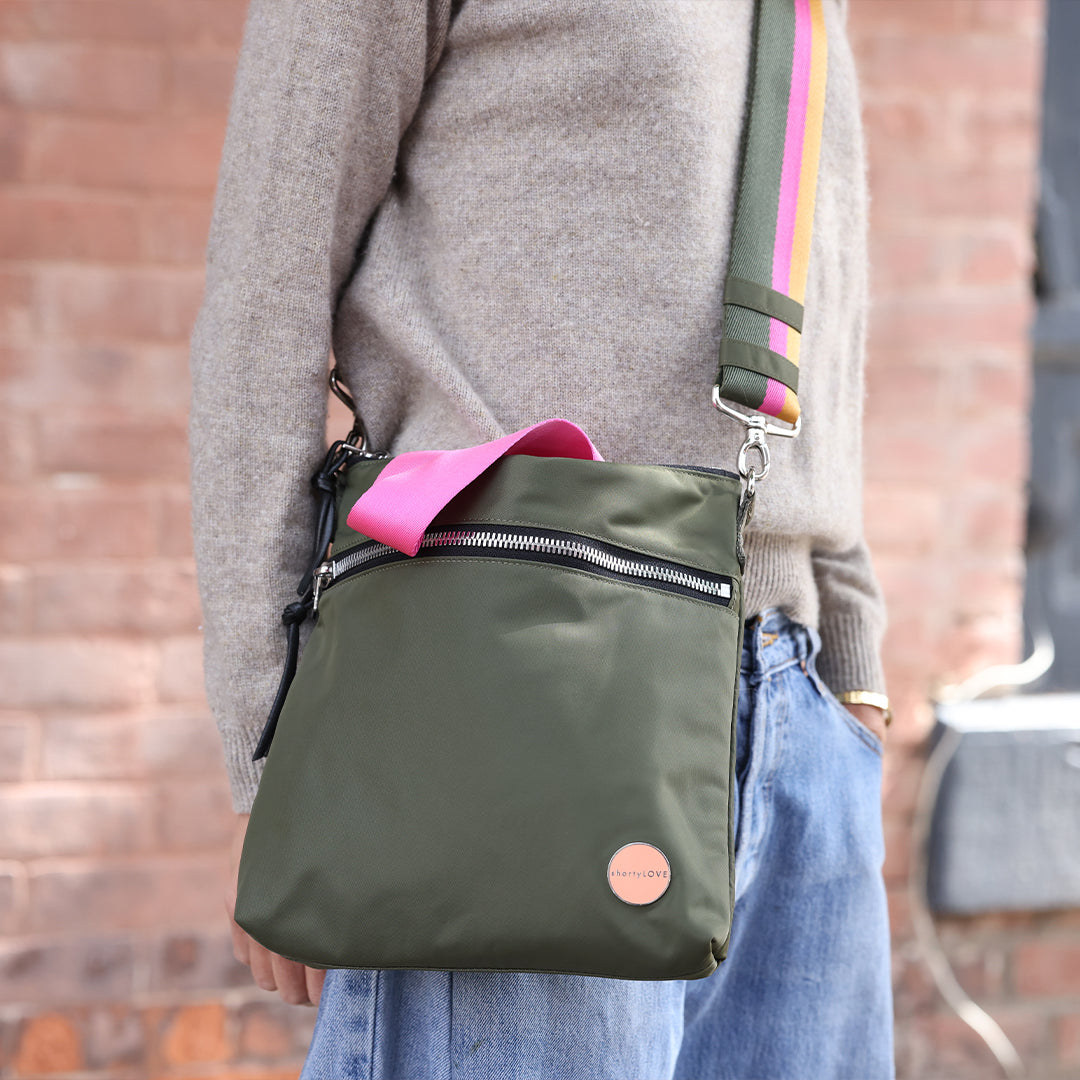 Person wearing an army green amuse crossbody bag with pink strap against a brick wall.