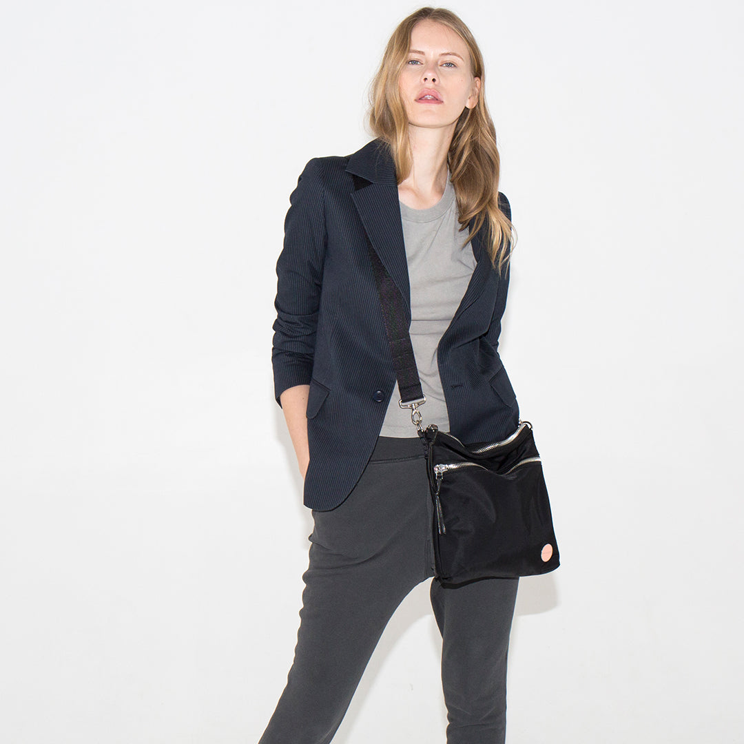 Woman wearing a navy blazer, gray top, and pants holding a black amuse crossbody handbag on a white background