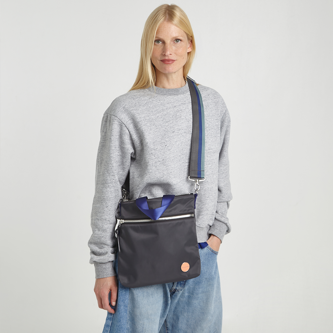 Woman wearing a gray sweatshirt and blue jeans with a charcoal amuse crossbody bag on a light gray background