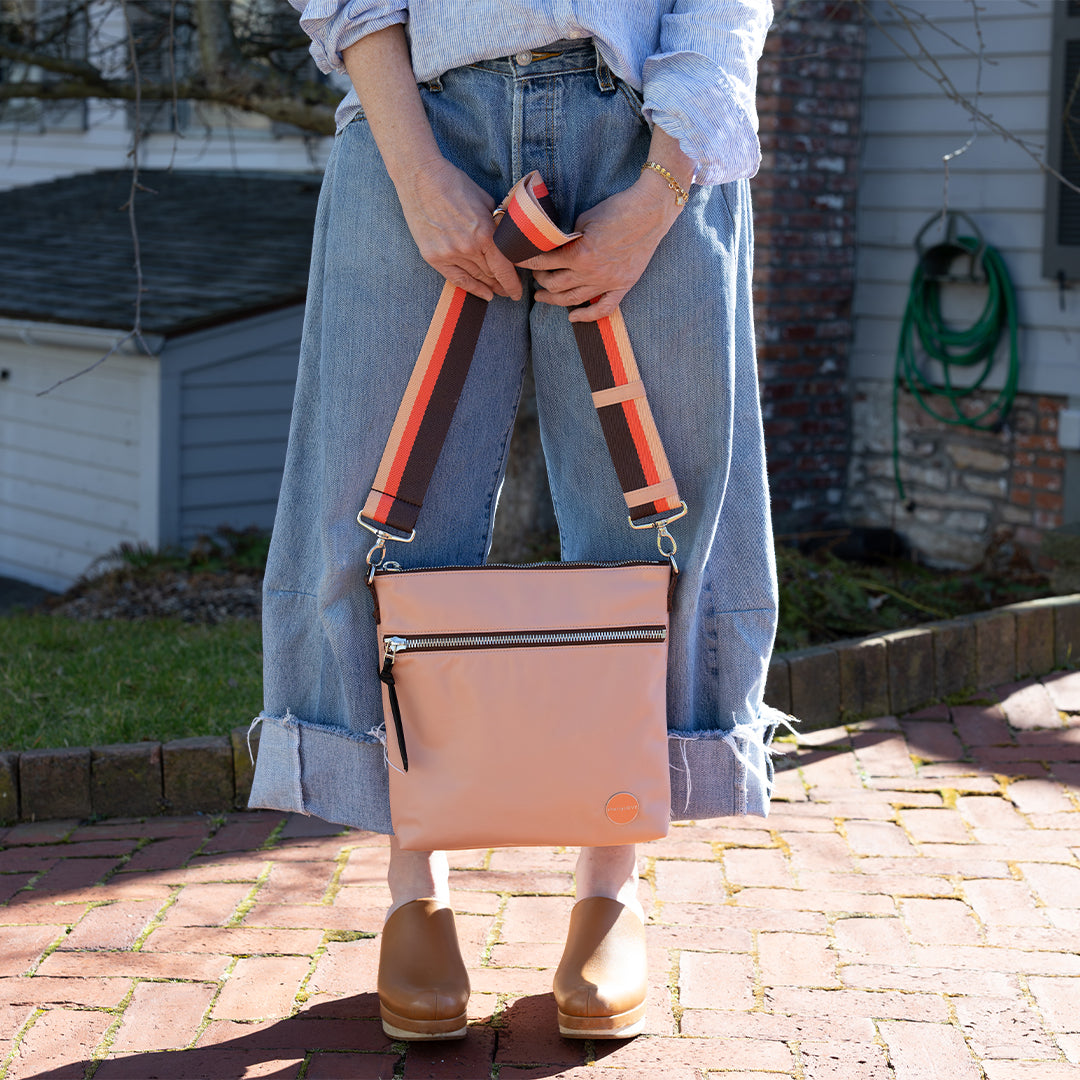 Person holding a dusty peach crossbody bag with striped handles on a brick patio.