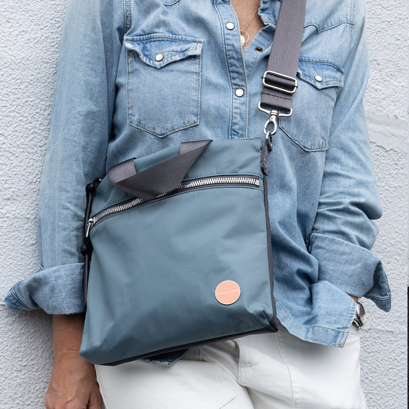 Person wearing a blue denim top with a steel blue amuse crossbody bag against a light gray wall.