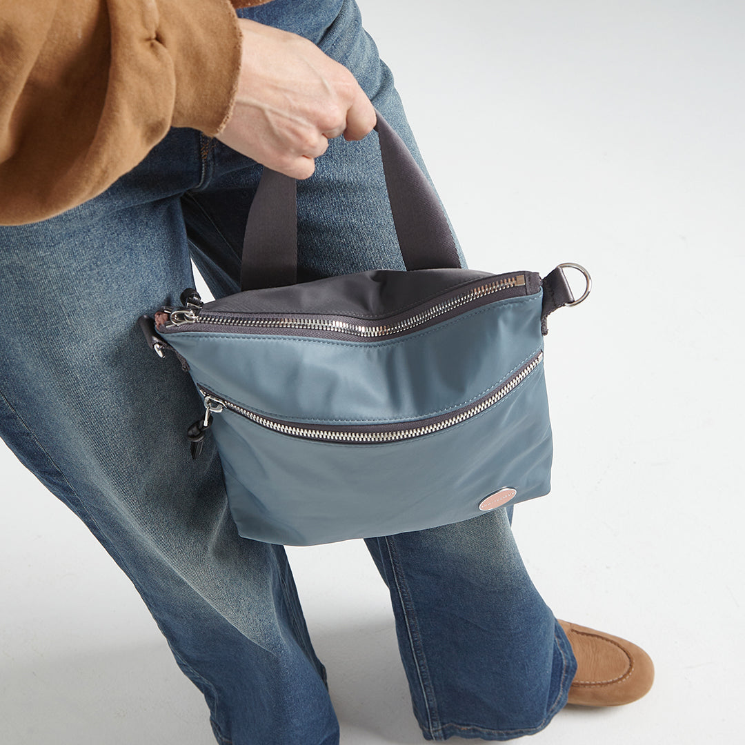 Person holding a steel blue amuse crossbody bag with zippers on a plain background