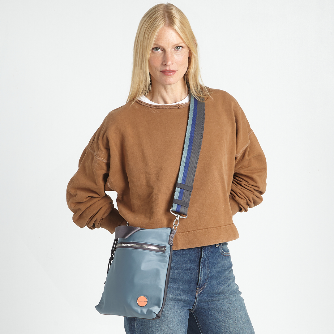 Woman wearing a brown sweatshirt and blue jeans with a steel blue crossbody bag with a strip[ed strap on a white background