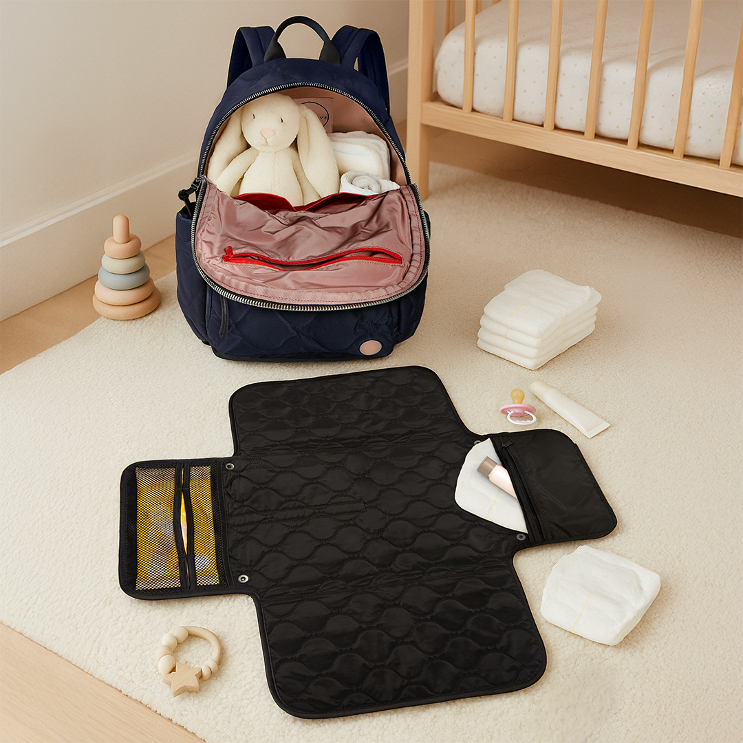 Navy blue backpack with baby items on a beige carpet with a black baby changing mat in a nursery.