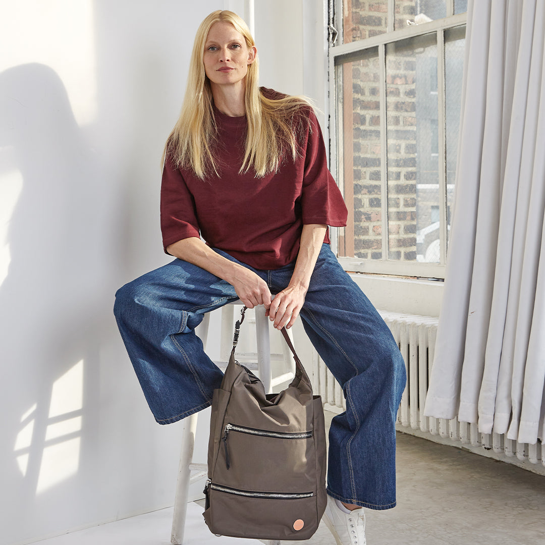 Woman sitting on a stool holding a mocha backpack in a bright room with large windows.