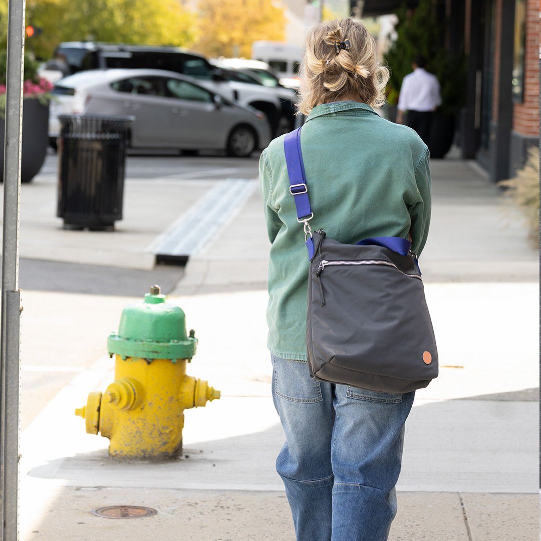 Person with a charcoal wonder crossbody bag standing next to a yellow fire hydrant on a sidewalk.
