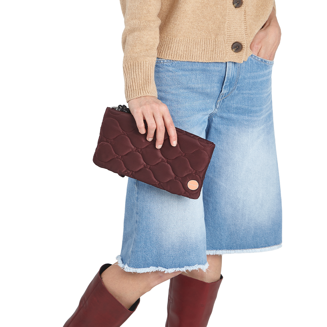 Person holding a bordeaux cleo quilted clutch with a beige sweater, blue denim skirt, and red boots on a white background 
