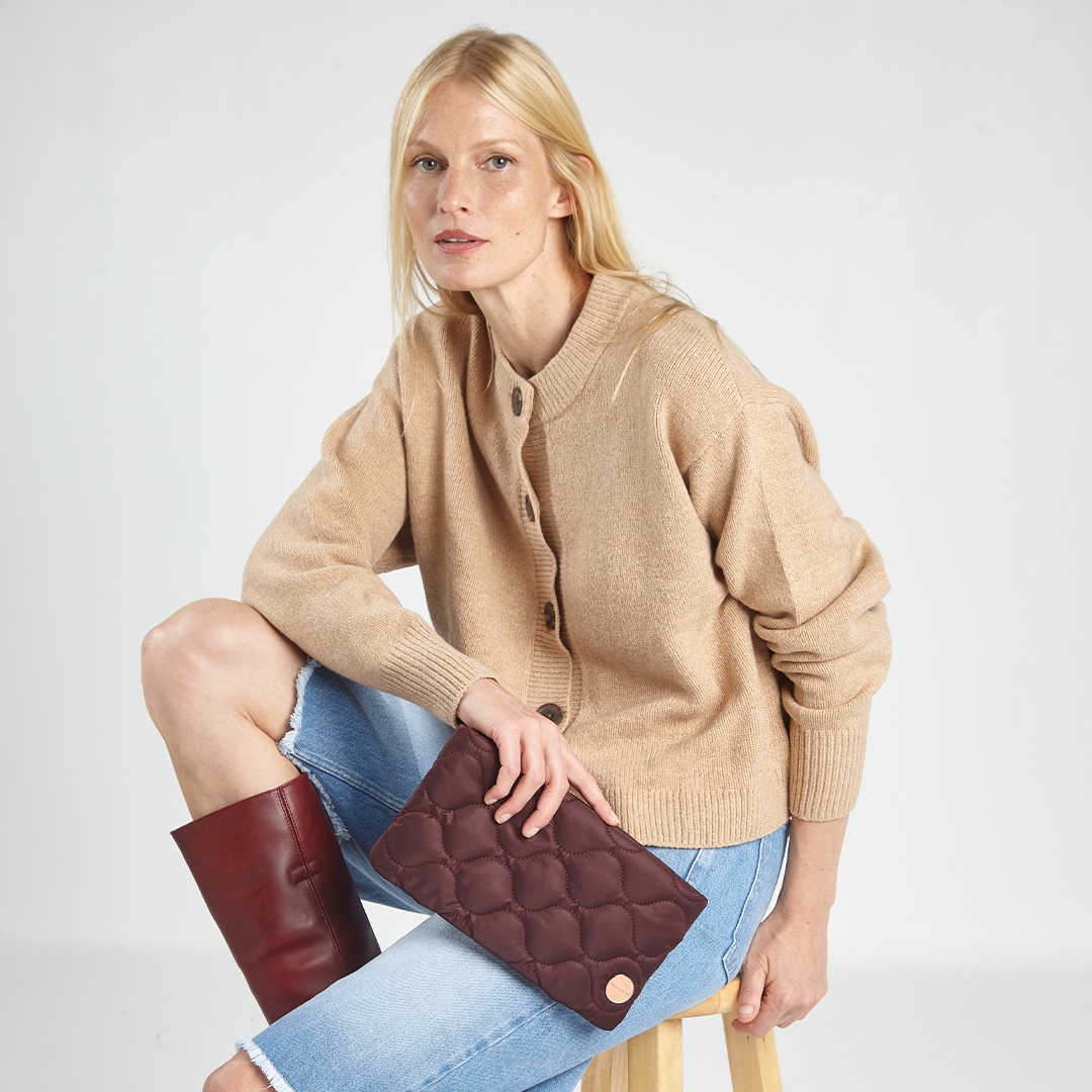 Woman wearing a beige cardigan and holding a bordeaux quilted clutch against a white background