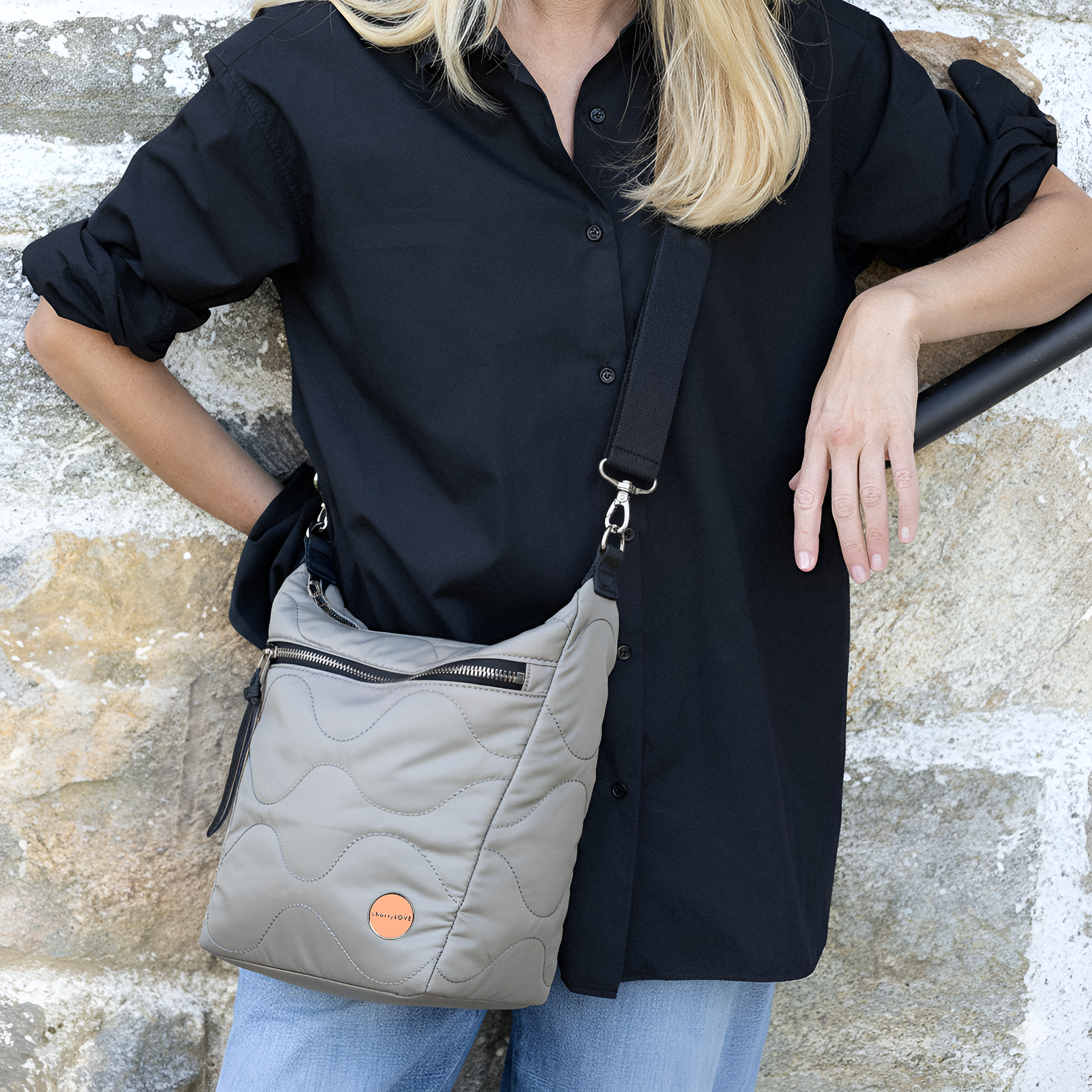 Person wearing a black shirt and holding a gray quilted marcel crossbody bag with a stone wall background
