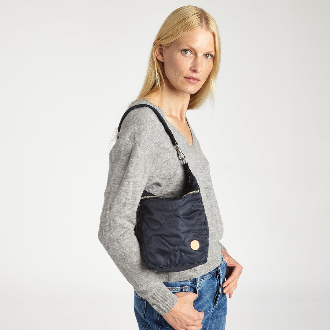 Woman wearing a gray sweater and blue jeans with a midnight blue marcel quilted crossbody bag on a white background