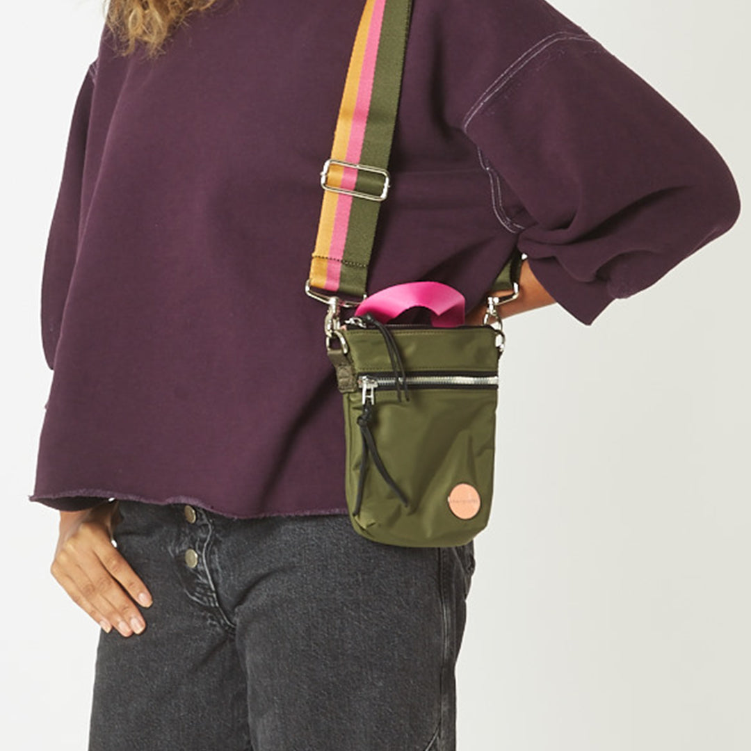Person wearing an army green shorthand crossbody bag with a colorful strap on a plain background