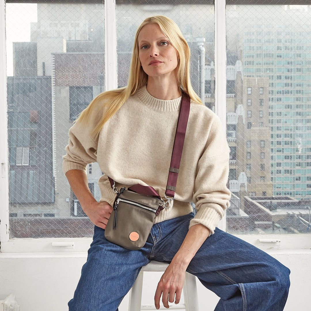 Woman sitting on a stool wearing a beige sweater and blue jeans, with a small mocha shorthand crossbody bag.