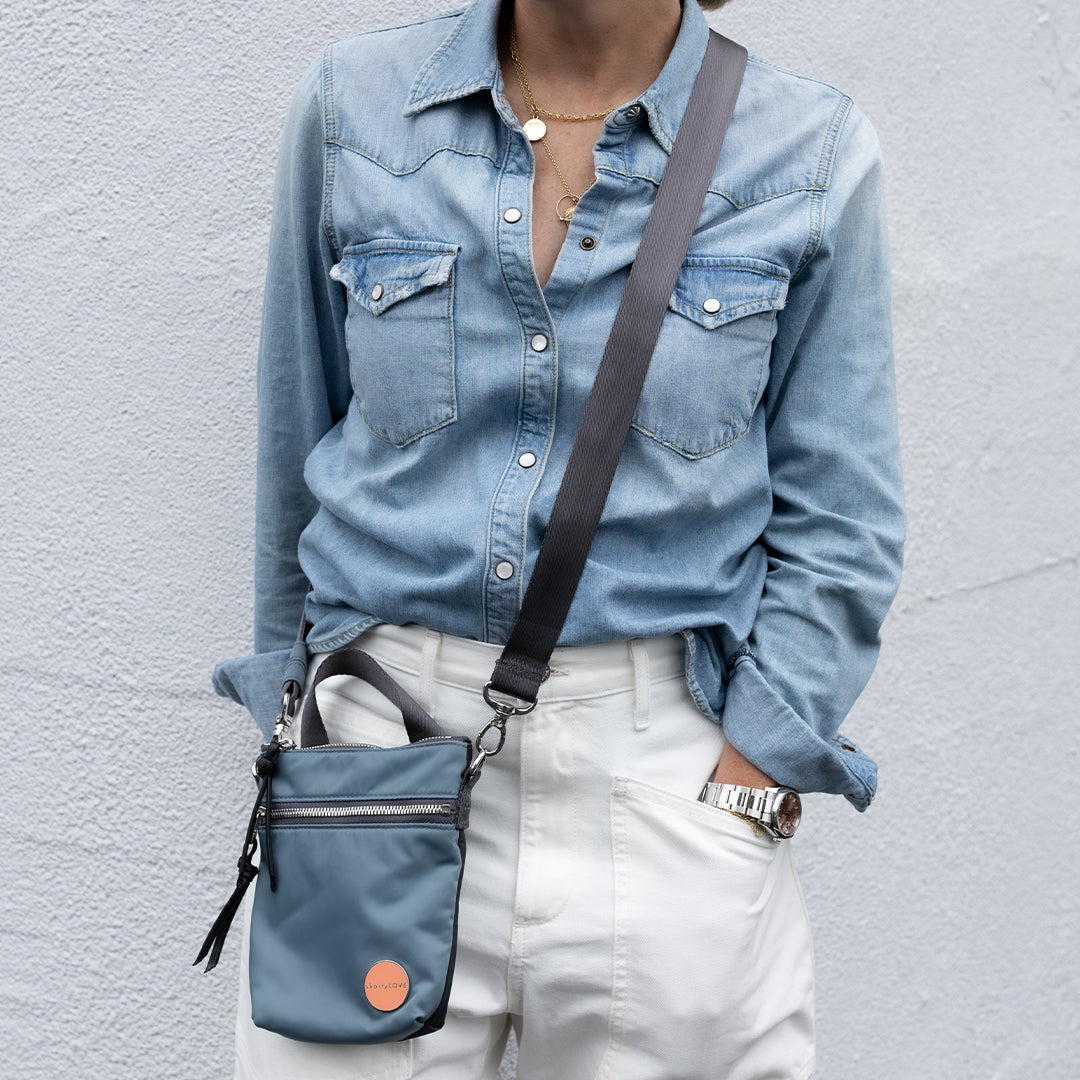 Person wearing a denim shirt and white pants with a steel blue/charcoal shorthand crossbody bag against a gray background