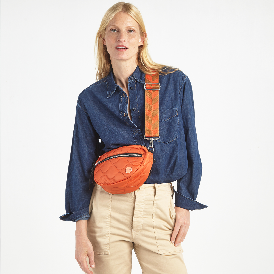 Woman wearing a blue denim shirt and beige pants with an orange crossbody bag on a white background